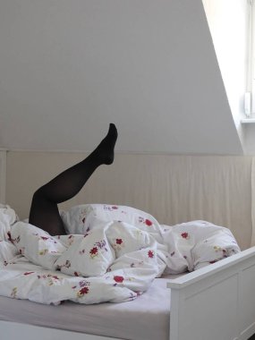 Mansard odasında ortaya çıkarılan beyaz yatakta battaniye altından yükselen siyah külotlu çorap seksi kadın bacak