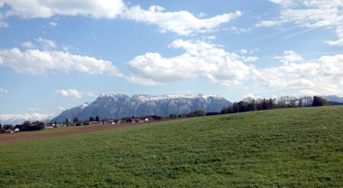 Parlak güneşli bir günde beyaz bulutlar ile mavi gökyüzü altında uzaklarda çiçek açan dağ çayırları ve yüksek kar tepesinde köy ile panoramik kırsal manzara