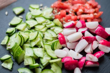 Dilimlenmiş taze salatalık, domates, kırmızı turp ve siyah bir arka plan üzerinde tohum. Sebzeden salata pişirmek. Sağlıklı beslenme kavramı, vejetaryenlik.