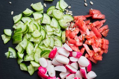 Dilimlenmiş taze salatalık, domates, kırmızı turp ve siyah bir arka plan üzerinde tohum. Sebzeden salata pişirmek. Sağlıklı beslenme kavramı, vejetaryenlik.