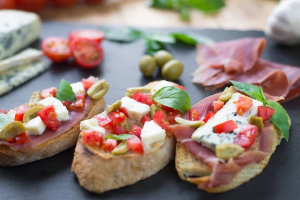 Tradiční italská brusšeta s modrým sýrem, Feta, rajčaty, lístky bazalky, Jamon na černém kamenném pozadí. — Stock fotografie