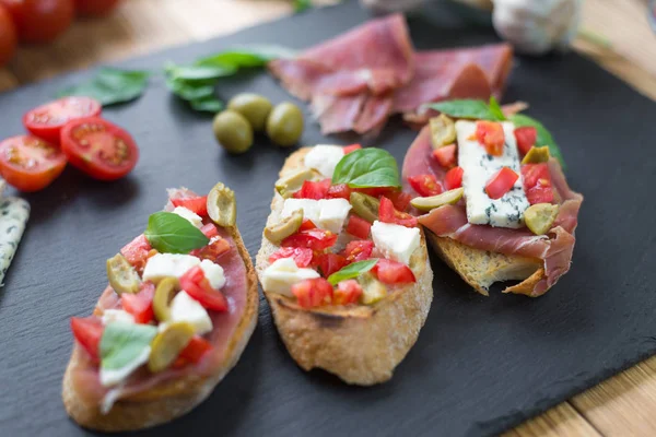 Bruschetta tradicional italiana con queso azul, feta, tomates, hojas de albahaca, jamón sobre fondo de piedra negra . —  Fotos de Stock