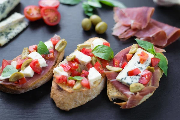 Bruschetta tradicional italiana con queso azul, feta, tomates, hojas de albahaca, jamón sobre fondo de piedra negra . — Foto de Stock