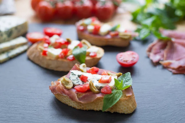 Bruschetta tradicional italiana con queso azul, feta, tomates, hojas de albahaca, jamón sobre fondo de piedra negra . —  Fotos de Stock