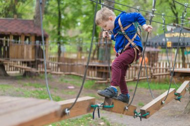 Küçük bir çocuk halat parkında eğitim göreyor. Çocuk engelli parkura tırmanıyor. Temiz havada parkta aktif rekreasyon.