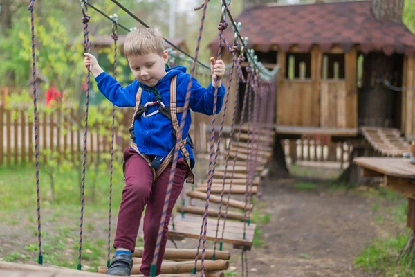 Um rapazinho está a treinar num parque de cordas. A criança sobe o curso de obstáculos. Recreação ativa no parque ao ar livre . — Fotografia de Stock