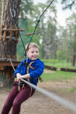 Küçük bir çocuk halat parkında eğitim göreyor. Çocuk engelli parkura tırmanıyor. Temiz havada parkta aktif rekreasyon.