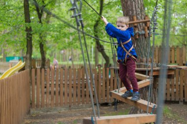 Küçük bir çocuk halat parkında eğitim göreyor. Çocuk engelli parkura tırmanıyor. Temiz havada parkta aktif rekreasyon.