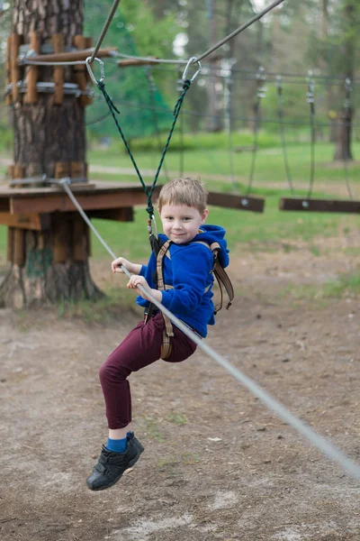 Um rapazinho está a treinar num parque de cordas. A criança sobe o curso de obstáculos. Recreação ativa no parque ao ar livre . — Fotografia de Stock