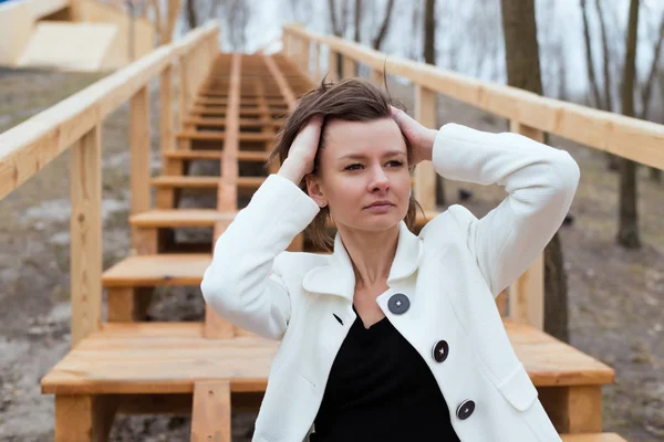 Dívka v bílém saku sedí na dřevěných schodech v parku. — Stock fotografie