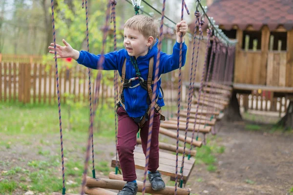 Um rapazinho está a treinar num parque de cordas. A criança sobe o curso de obstáculos. Recreação ativa no parque ao ar livre . — Fotografia de Stock
