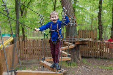 Küçük bir çocuk halat parkında eğitim göreyor. Çocuk engelli parkura tırmanıyor. Temiz havada parkta aktif rekreasyon.