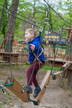 Küçük bir çocuk halat parkında eğitim göreyor. Çocuk engelli parkura tırmanıyor. Temiz havada parkta aktif rekreasyon.