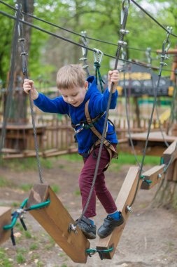 Küçük bir çocuk halat parkında eğitim göreyor. Çocuk engelli parkura tırmanıyor. Temiz havada parkta aktif rekreasyon.
