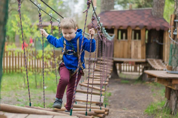 Um rapazinho está a treinar num parque de cordas. A criança sobe o curso de obstáculos. Recreação ativa no parque ao ar livre . — Fotografia de Stock