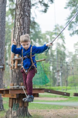 Küçük bir çocuk halat parkında eğitim göreyor. Çocuk engelli parkura tırmanıyor. Temiz havada parkta aktif rekreasyon.