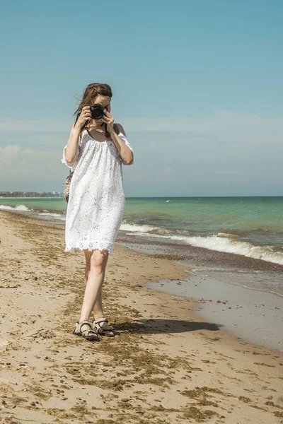 Kvinna fotograf vid havet. En medelålders kvinna i en vit klänning med en kamera går längs kusten. Sommar turism, fotografering sevärdheter på semester. — Stockfoto