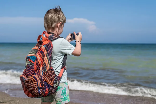 Мальчик-фотограф. Ребенок с рюкзаком делает фото на пляже . — стоковое фото
