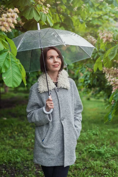 Vrouw in een jas op de achtergrond van groene bomen onder een paraplu tijdens de regen. — Stockfoto