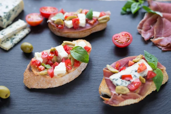 Bruschetta tradicional italiana con queso azul, feta, tomates, hojas de albahaca, jamón sobre fondo de piedra negra . —  Fotos de Stock