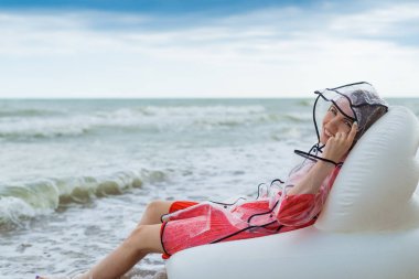 Memnun, neşeli bir kadın yağmur sırasında yağmurluk içinde sahilde dinleniyor.