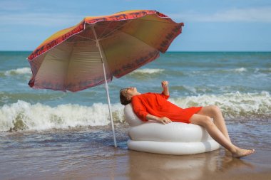 Bir kadın güneş şemsiyesi altında suda şişme bir sandalyede otururken denizde dinleniyor.