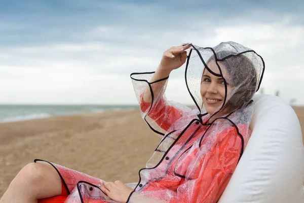 Şeffaf yağmurluk lu bir kadın hafif bir yağmur sırasında deniz kenarında şişme bir sandalyede, okyanusta rahatlar..