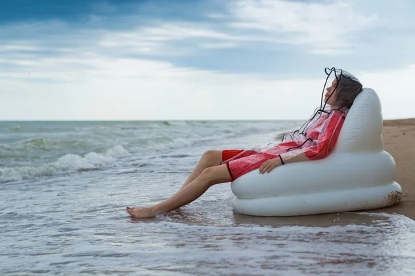 A contented, joyful woman rests on the beach in a raincoat during rain.