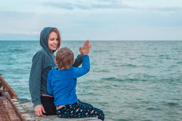 माँ और उसका छोटा बेटा समुद्र, महासागर पर बंदरगाह पर एक साथ बैठे हैं और खुशी से एक दूसरे के हाथ clapping . — स्टॉक फ़ोटो, इमेज