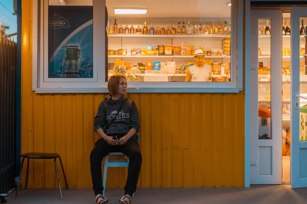 Gribovka, Odessa region- June 07, 2019: A middle-aged woman sits on a chair near a small shop with food and alcohol in the evening. — Stock Photo, Image