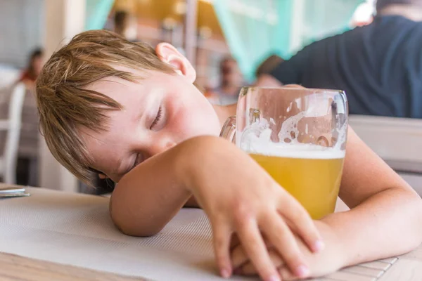 The little boy fell asleep in front of a glass of beer at the table.