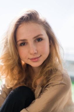 Kıvırcık kahverengi saçlı genç bir kız kameraya bakıyor.