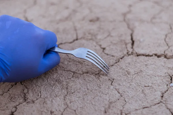 Het concept van voedseltekorten, milieurampen. Een rubberen handschoenen houdt een vork op de achtergrond van gebarsten droge grond. — Stockfoto