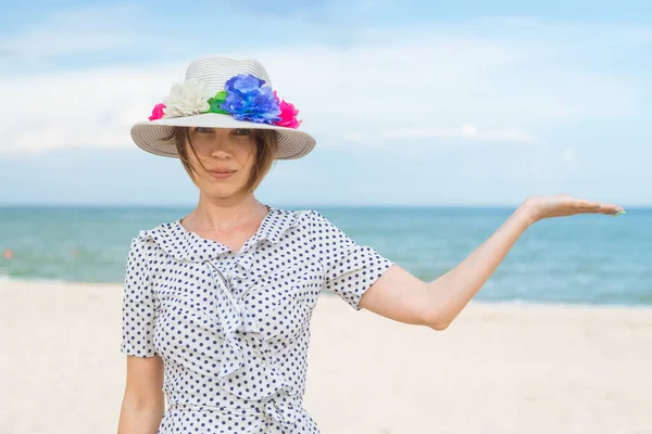 A middle-aged woman in a hat and dress on the seashore, the ocean holds her hand palm up. Place for text. Leisure, vacation, serenity, freedom concept.