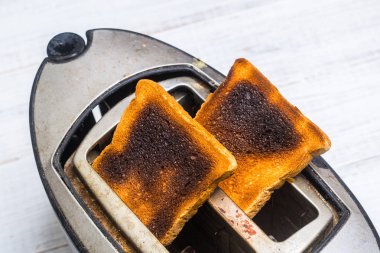 Beyaz ahşap arka planda yanmış, abartılı ekmek ile kirli eski tost makinesi. Kırık, arızalı mutfak aletleri.