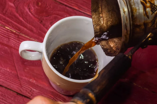 El café se vierte de un cezve en una taza sobre un fondo de madera . — Foto de Stock