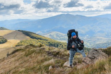 Orta yaşlı bir adam büyük bir sırt çantasıyla dağlarda yürüyüş yapıyor..