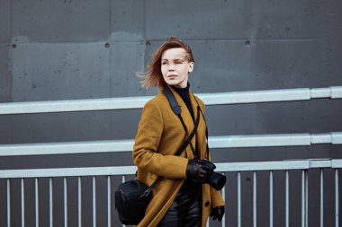 Gri arka planda elinde kamera olan ceketli bir kadın. Gezgin, turist.