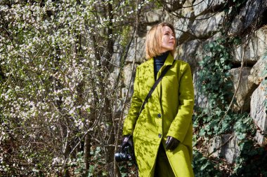 Yeşil ağaçta kamerası olan ceketli bir kadın..