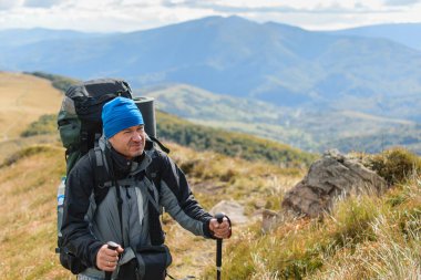 Orta yaşlı bir adam büyük bir sırt çantasıyla dağlarda yürüyüş yapıyor..
