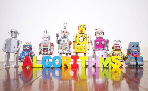 ряд ретро-роботов на деревянном полу со словом ALGORITHM
