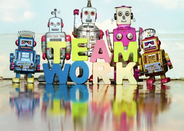 the word TEAM WORK  with wooden letters and 5 retro bots on a wooden floor say hi