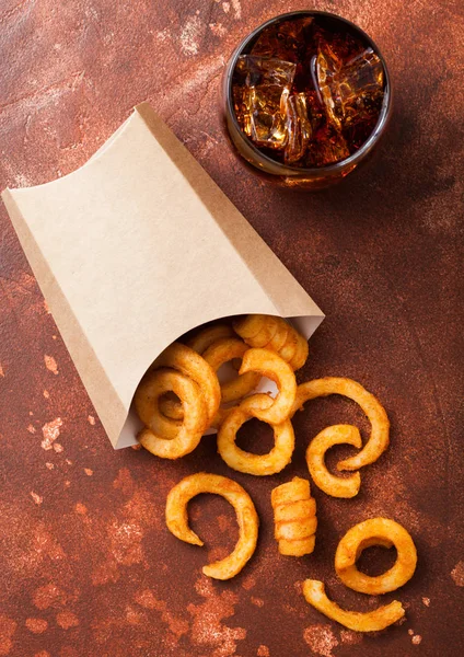 Curly fries fast food snack in paper container with glass of cola on rusty kitchen background. Unhealthy junk food