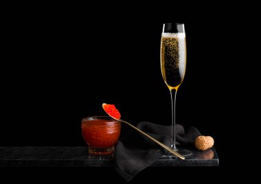 Altın kaşıkta kırmızı havyar ve siyah arka planda mermer panoda cam havyar dolu zarif sarı şampanya kadehi.