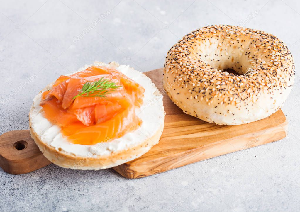 Fresh healthy bagel sandwich with salmon, ricotta and dill on vintage chopping board on light kitchen table background.