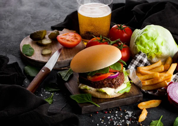 Fresh beef burger with sauce and vegetables and glass of lager craft beer with potato chips fries on stone kitchen background.