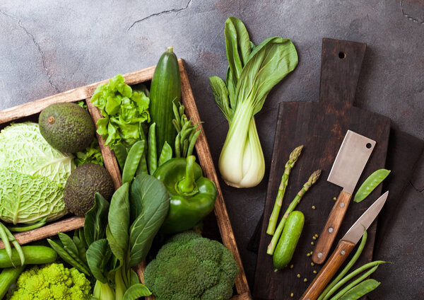 Assorted green toned raw organic vegetables in wooden box on dark stone background. Avocado, cabbage, cauliflower and cucumber with trimmed beans and chopping board with knife.