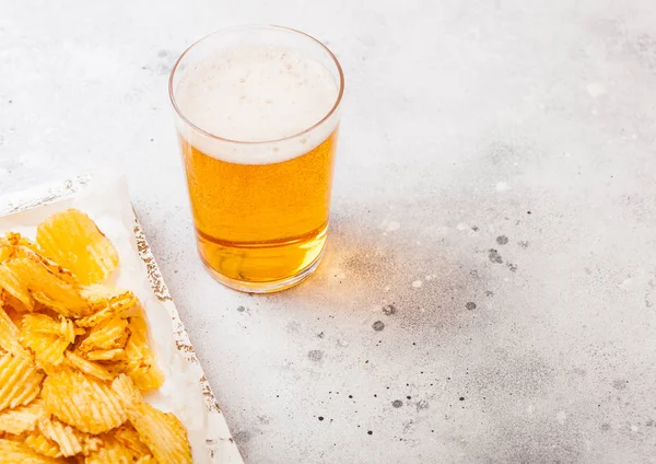 Glass of craft lager beer with potato crisps snack in vintage wooden box and opener on stone kitchen background. Beer and snack.