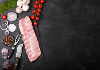 Siyah taş zemin üzerine kurulu ve vintage et çatalı ve bıçağı doğrama üzerinde taze çiğ Domuz kaburga. Taze domates ve kırmızı soğan sarımsak, tuz ve biber ile.