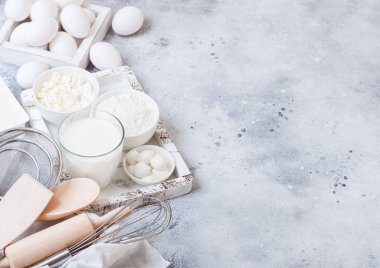 Beyaz masa arka planında taze süt ürünleri. Bir bardak süt, bir kase un, süzme peynir ve yumurta. Bir kutu kap kacak. Kaliteli ahşap kutuda çırpma ve spatula. Üst manzara.. 